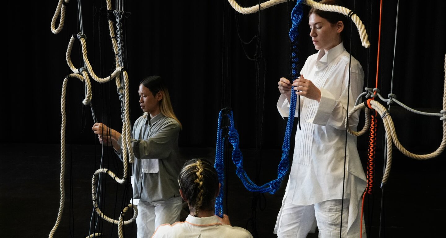 Auf dem Bild sind vor schwarzem Hintergrund die Performer:innen Duc, Emmilou und Lau. Sie tragen helle Kleidung und knoten an einem Seilkonstrukt, bestehend aus einer Struktur von verschiedenen Seilen. Darin verknotet ist ein orangefarbener, ein blauer und ein grauer Knotenstrang. Duc knotet links in der Hocke auf den Knien an grauen Seilen, Emmilou steht rechts im Bild und verknotet den blauen Strang. Lau ist uns im Bildvordergrund mit dem Rücken zugewandt und führt das Knoten des blauen Stranges fort.